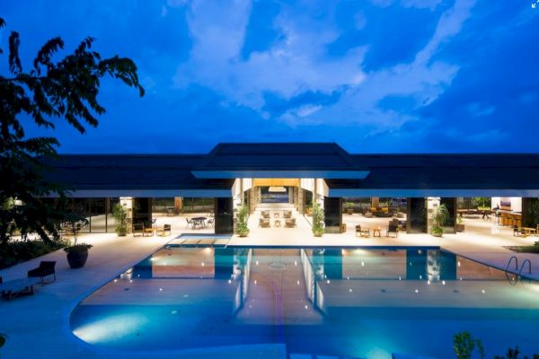 A modern building with large windows lights up against a twilight sky, reflecting in a spacious swimming pool in the foreground.