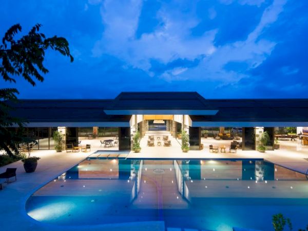A modern building with large windows lights up against a twilight sky, reflecting in a spacious swimming pool in the foreground.