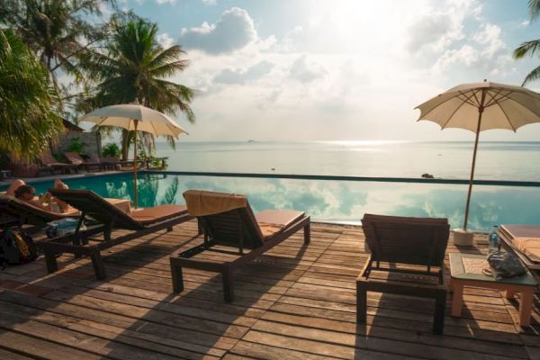 A scenic poolside view with lounge chairs, umbrellas, palm trees, and a calm ocean under a partly cloudy sky.