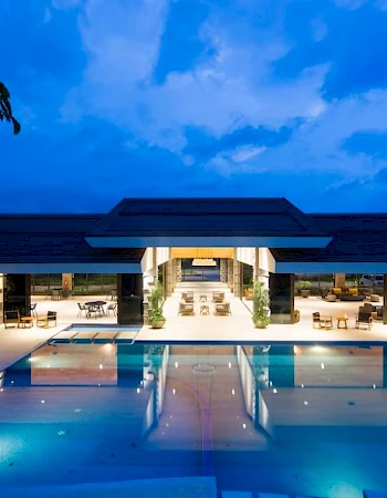 A luxurious modern building with a large illuminated pool, surrounded by outdoor seating, under a twilight sky.