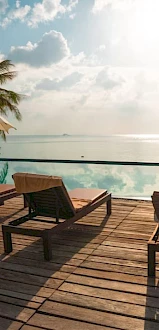 A wooden deck with lounge chairs and umbrellas overlooks an infinity pool facing the ocean, under a partly cloudy sky.