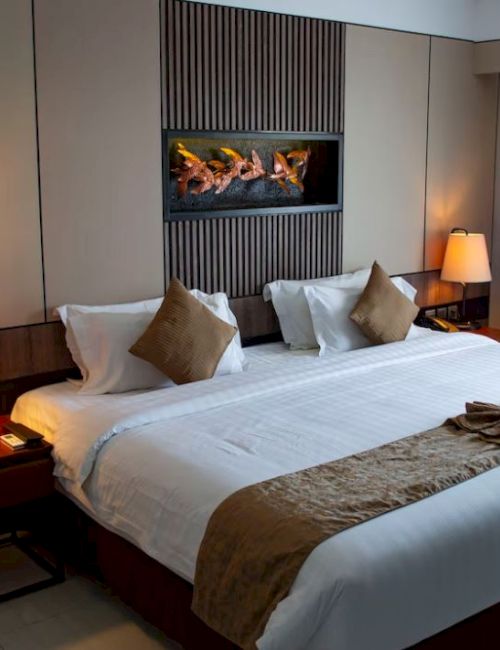 A neatly made hotel room with a large bed, pillows, lamps, and a decorative inset feature above the bed. Curtains are drawn to one side.