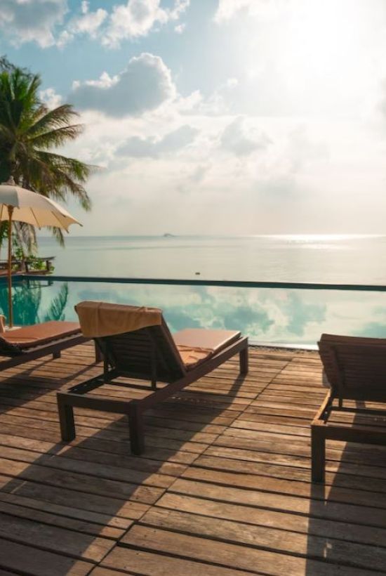 A serene poolside scene with sun loungers, umbrellas, and palm trees overlooking a tranquil sea under a partly cloudy sky.