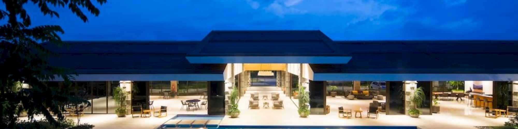A luxurious modern building with a large outdoor pool is lit up against a twilight sky, creating a serene and inviting ambiance.