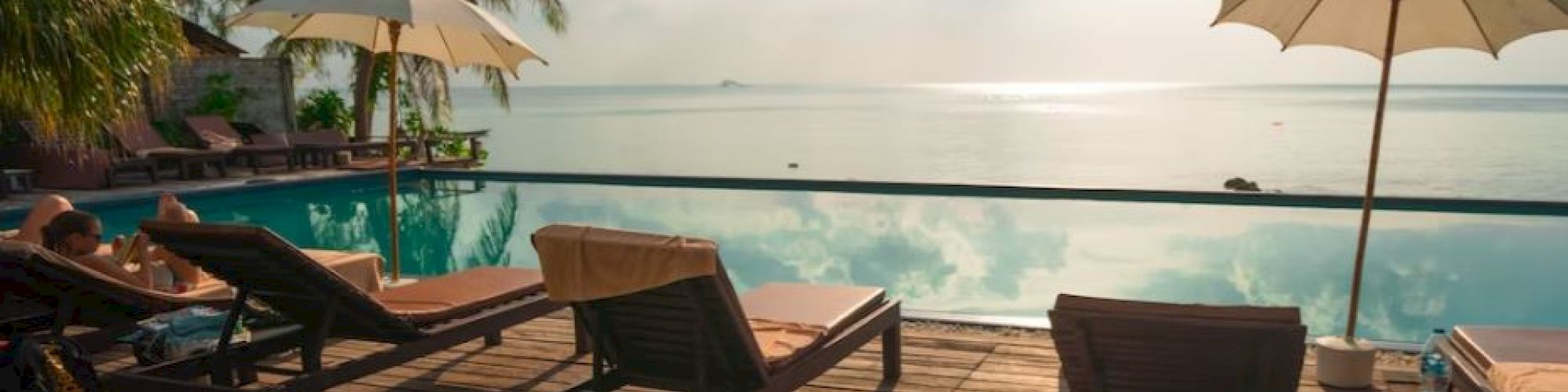 A scenic view of a tropical poolside with sun loungers, umbrellas, palm trees, and a tranquil ocean backdrop under a bright sky.