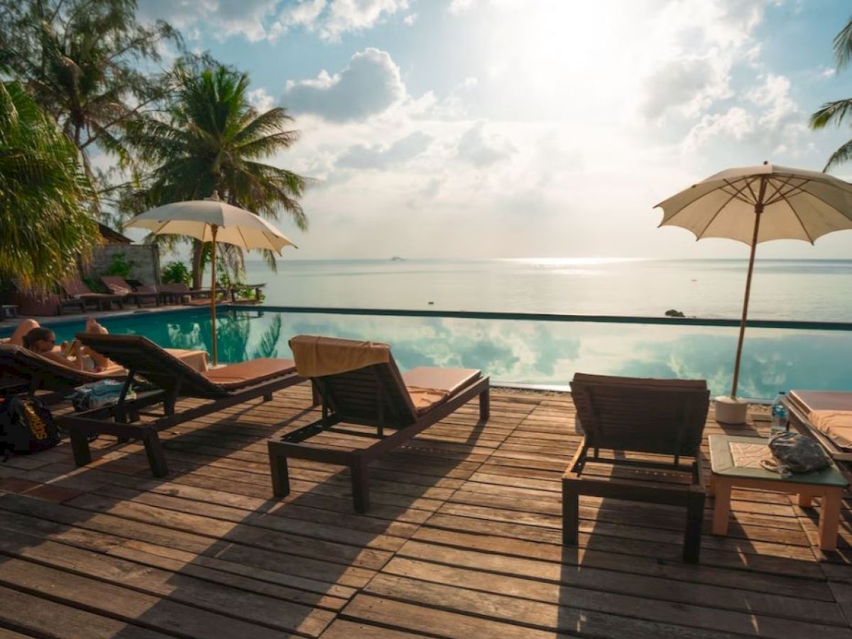 A tranquil poolside scene with lounge chairs, umbrellas, and palm trees overlooking serene water under a partly cloudy sky.