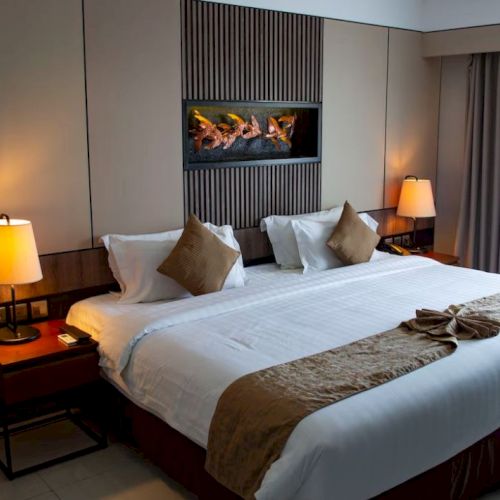 A cozy hotel room with a large bed, two side tables with lamps, decorative pillows, and art on the wall; curtains partially drawn.