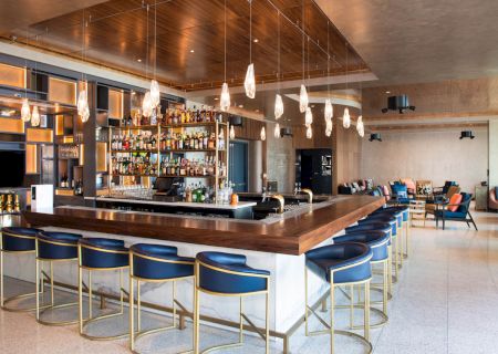 A modern bar with blue stools, overhead lighting, and a well-stocked counter surrounded by seating in an open, stylish space.