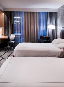 A modern hotel room with two beds, a desk, a TV, and stylish decor, featuring a patterned accent wall and floor-to-ceiling curtains.