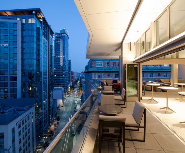 A modern balcony overlooking a city street with high-rise buildings, furnished with tables and chairs, and featuring large windows.