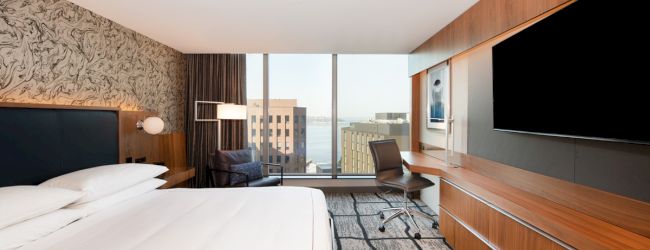 A modern hotel room features a large bed, desk, chair, and TV. The window offers a cityscape view with buildings and water in the distance.