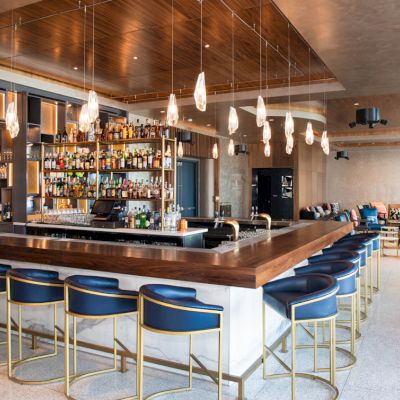 A modern bar with a wooden countertop, blue cushioned bar stools, pendant lighting, and a well-stocked back bar.