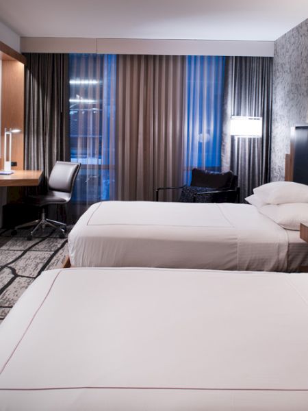 The image shows a modern hotel room with two beds, a large TV, a desk, a chair, and stylish lighting, creating a cozy atmosphere.