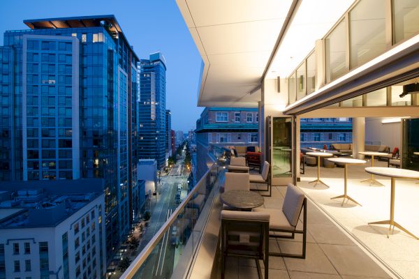 A modern cityscape at dusk with high-rise buildings, a sleek balcony, outdoor seating, glass walls, and illuminated interiors.