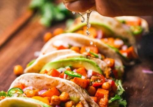 A hand squeezes lime over a row of tacos filled with vegetables and chickpeas on a wooden surface, garnished with lime and cilantro.