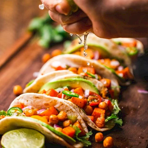 A hand squeezes lime over a row of tacos filled with vegetables and chickpeas on a wooden surface, garnished with lime and cilantro.