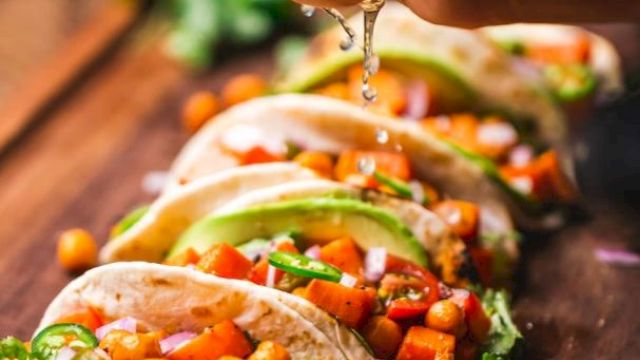 A hand squeezes lime over a row of tacos filled with vegetables and chickpeas on a wooden surface, garnished with lime and cilantro.