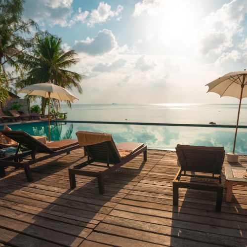 The image shows a serene poolside scene with lounge chairs, parasols, and a view of a calm sea under a partly cloudy sky.