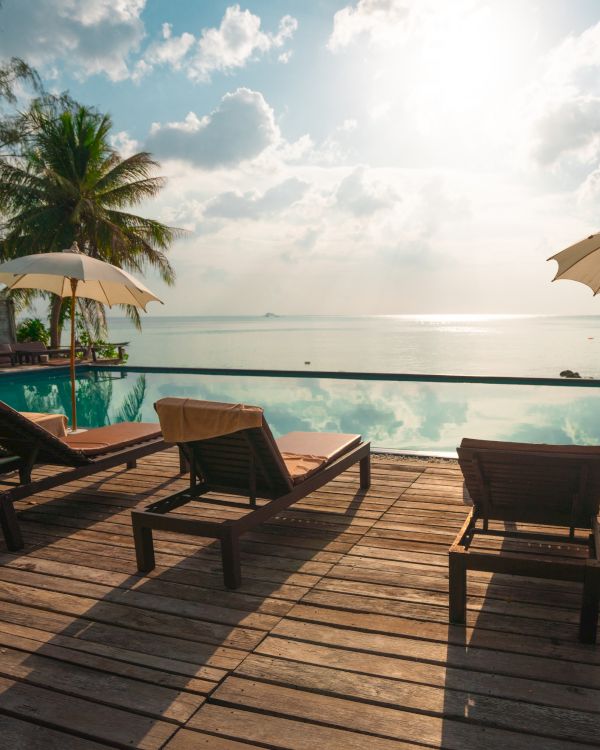 The image shows a serene poolside scene with lounge chairs, parasols, and a view of a calm sea under a partly cloudy sky.