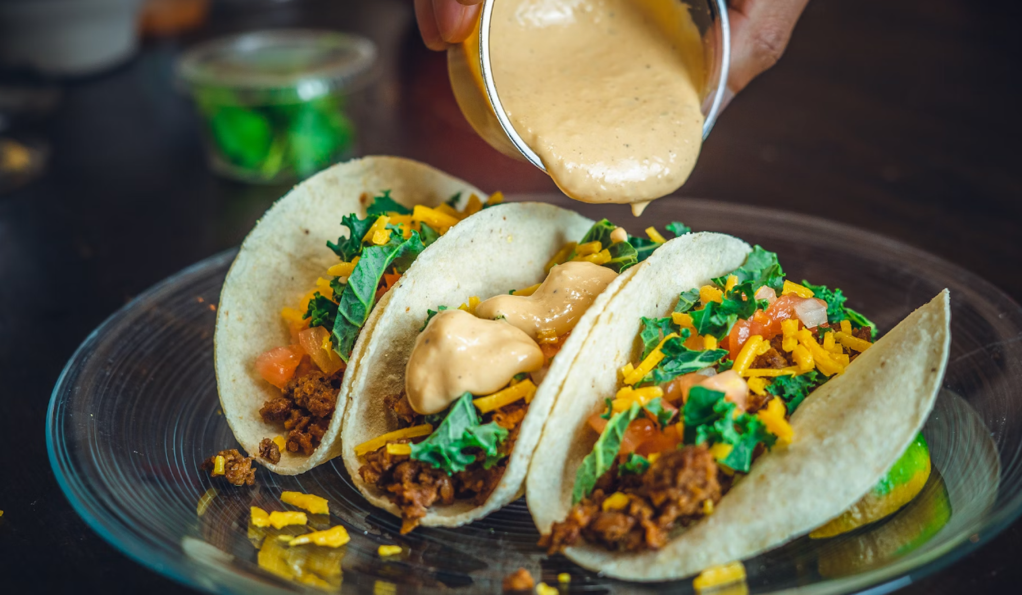 Three tacos with lettuce, tomato, cheese, and sauce being poured on top are arranged on a glass plate.
