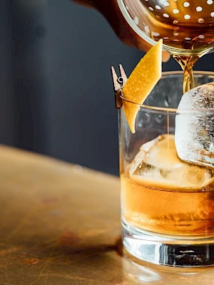 A cocktail is being poured into a glass with a large ice cube and a twist of citrus peel as garnish.