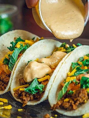Three tacos filled with meat, cheese, lettuce, and tomatoes, being topped with a creamy sauce on a plate.