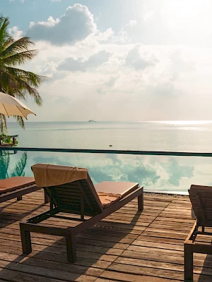 Lounge chairs and umbrellas by an infinity pool overlooking a calm sea under a partly cloudy sky, surrounded by palm trees.