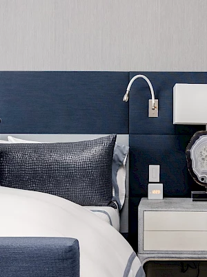 A modern bedroom with a navy and white color scheme, featuring a bed, pillows, wall light, and a nightstand with a lamp and decorative items.