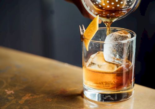A cocktail being poured into a glass with ice and a citrus twist garnish, placed on a wooden surface.