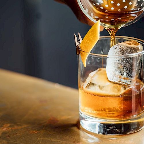 A cocktail being poured into a glass with ice and a citrus twist garnish, placed on a wooden surface.