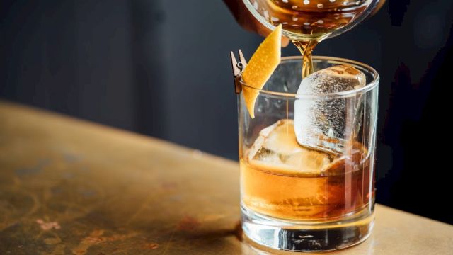 A cocktail being poured into a glass with ice and a citrus twist garnish, placed on a wooden surface.