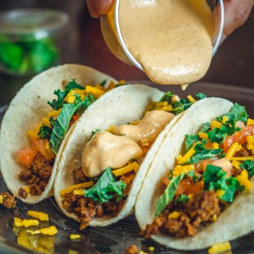 Three tacos filled with ground meat, cheese, tomatoes, and greens are being topped with a creamy sauce on a plate.
