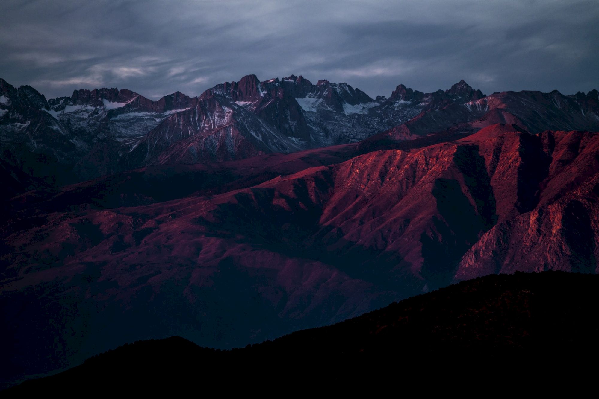 Mountains at dusk are bathed in red and purple hues under a cloudy sky, creating a dramatic and serene landscape.