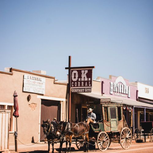 A vintage Western scene with a horse-drawn carriage in front of rustic buildings, including one labeled "O.K. Corral," under a clear blue sky.