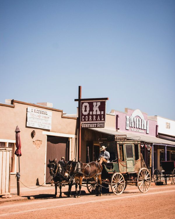 A vintage Western scene with a horse-drawn carriage in front of rustic buildings, including one labeled "O.K. Corral," under a clear blue sky.