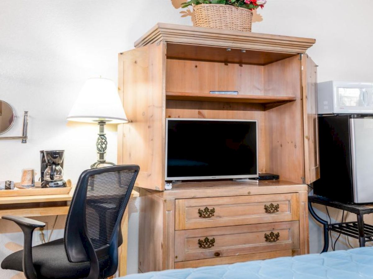 Room with a desk, chair, lamp, coffee maker, wooden cabinet with a TV, microwave, fridge, and a basket with flowers on top.