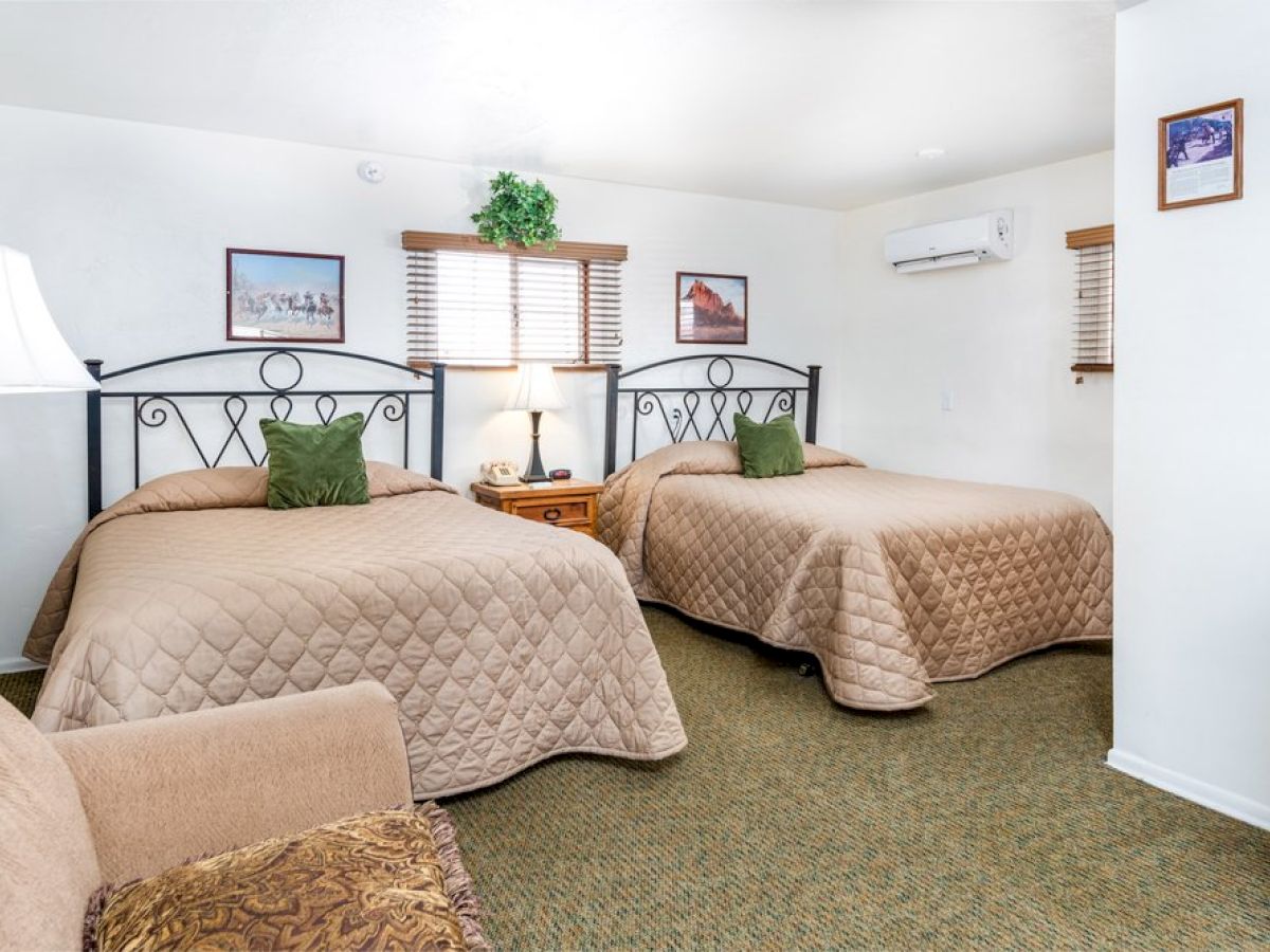 The image shows a cozy bedroom with two beds, lamps, a sofa, wall art, and carpeted floor, creating a welcoming atmosphere.