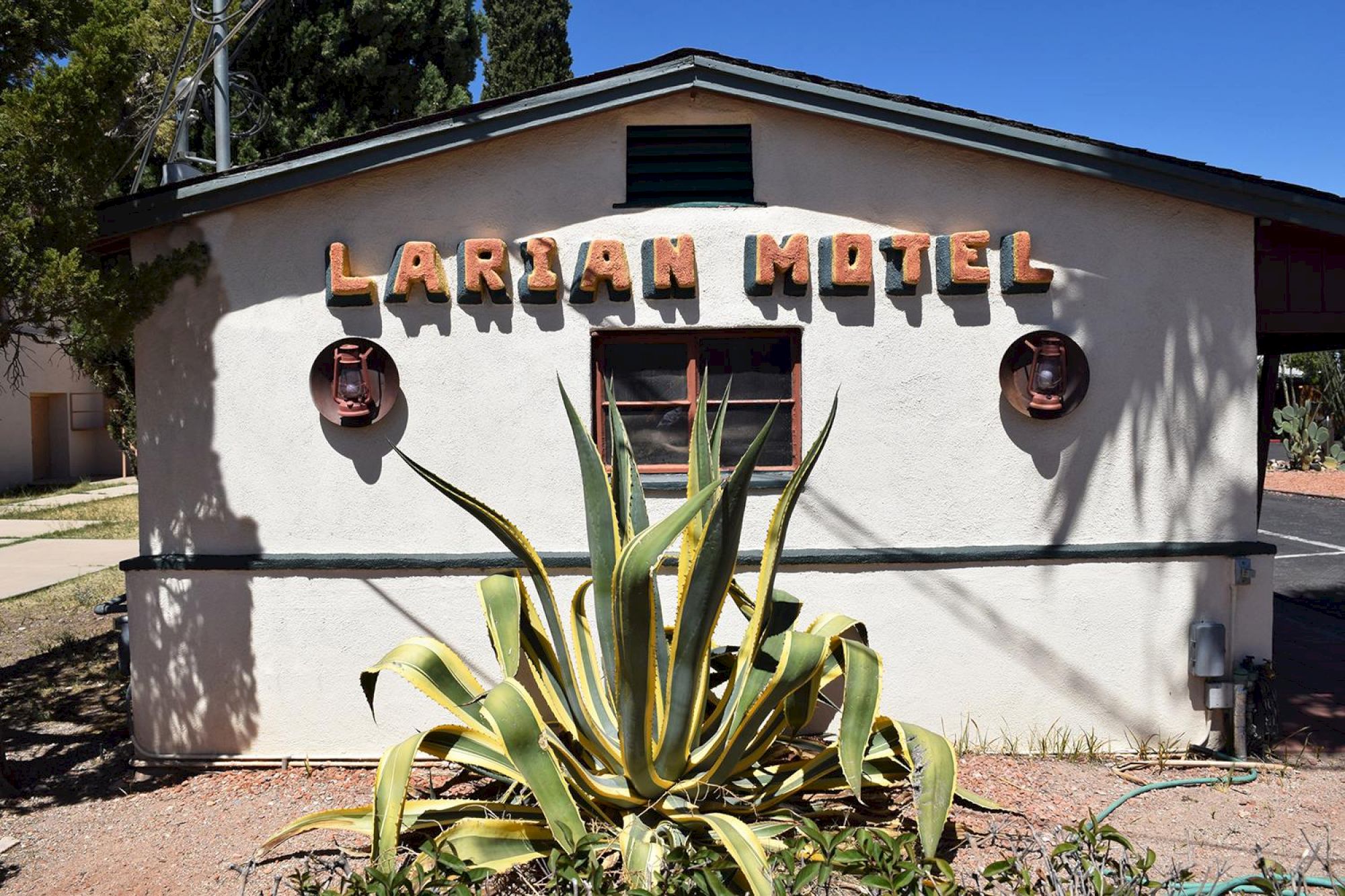 The image shows a building with a sign reading "Larian Motel," featuring desert plants in the foreground and a simple, rustic design.