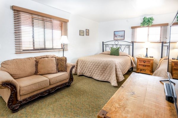 A cozy room with a bed, a beige sofa, wooden nightstands, and lamps. There's artwork on the walls and a TV on a wooden dresser.