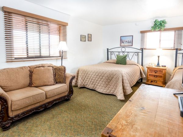 A cozy room with a bed, a beige sofa, wooden nightstands, and lamps. There's artwork on the walls and a TV on a wooden dresser.