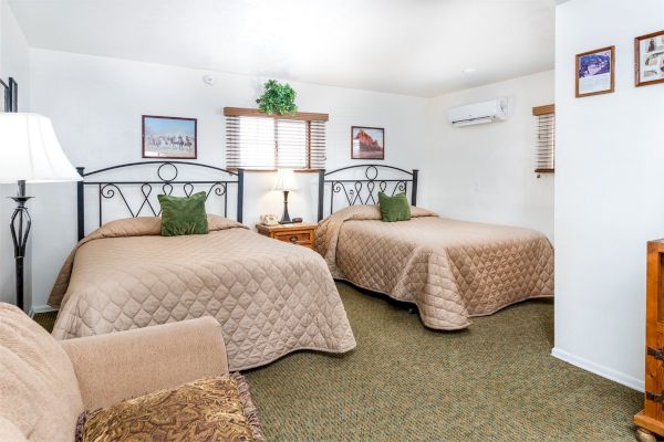 A cozy hotel room with two beds, a nightstand, and a table lamp. There's wall art and an AC unit. Green pillows accent beige bedding.