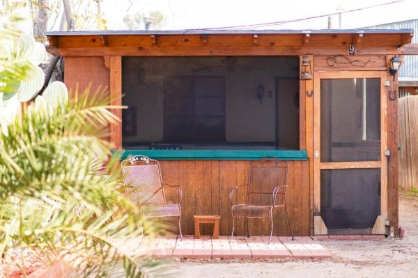 A small rustic cabin with a screened porch and two metal chairs outside on a brick pathway.
