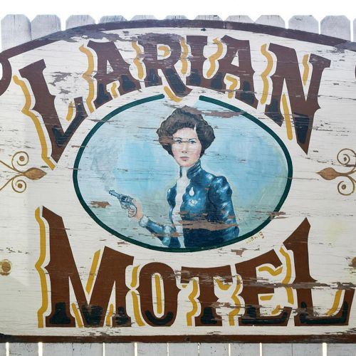 A vintage wooden sign with "Larian Motel" and a painted figure of a woman holding a gun in the center, set against a white fence.