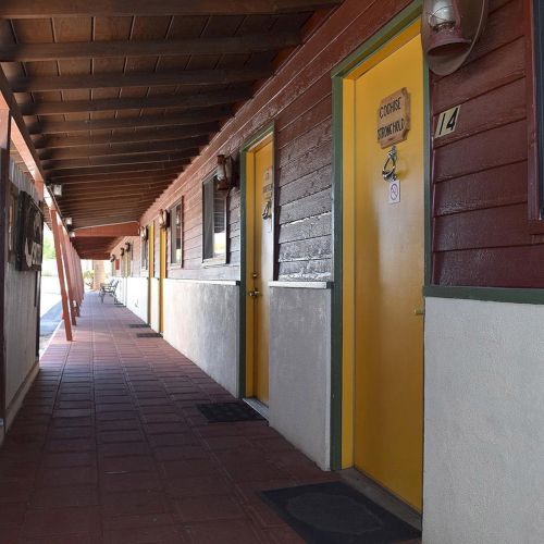 A long outdoor corridor with red and beige walls, yellow doors, and a tiled floor. Each door has a sign and a window nearby.