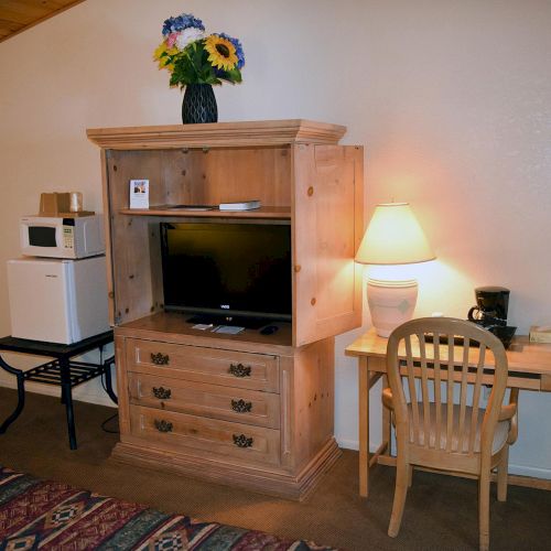 The image shows a hotel room corner with a TV, dresser, desk, lamp, chair, microwave, mini-fridge, and flower vase.