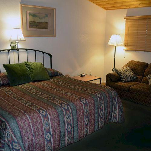 A cozy room with a bed, patterned bedspread, green pillows, an armchair, striped cushions, a lamp, and a framed picture on the wall.