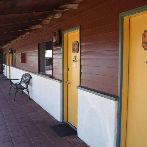 A motel exterior with a shaded porch, yellow doors labeled "Curly Bill" and "Luke Short," along with a bench under a rustic roof structure.