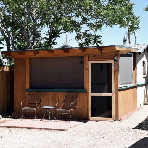 A small wooden building with a patio, two chairs, table, and surrounded by trees and fences under a clear sky.