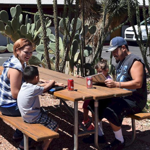 A family sits at a picnic table outdoors, enjoying drinks and snacks, surrounded by desert plants and cacti in the background.