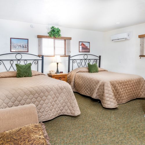 The image shows a cozy hotel room with two beds, wall art, a lamp on a nightstand, and a small air conditioning unit mounted on the wall.